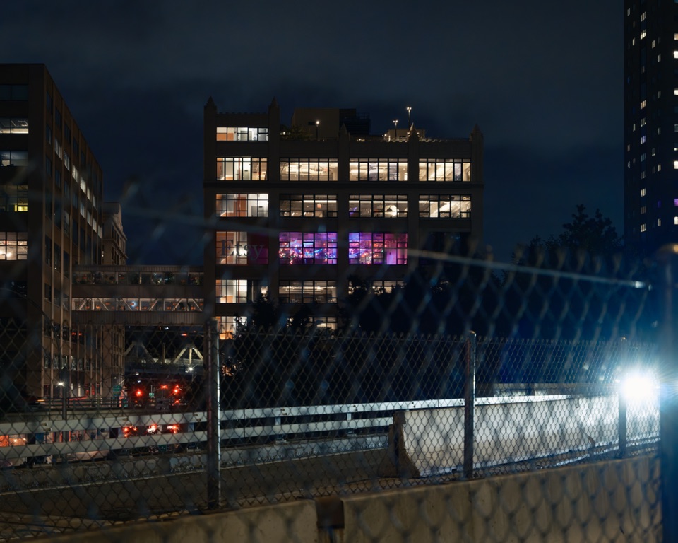 A party in a lit building, seen past a chain-link fence at night