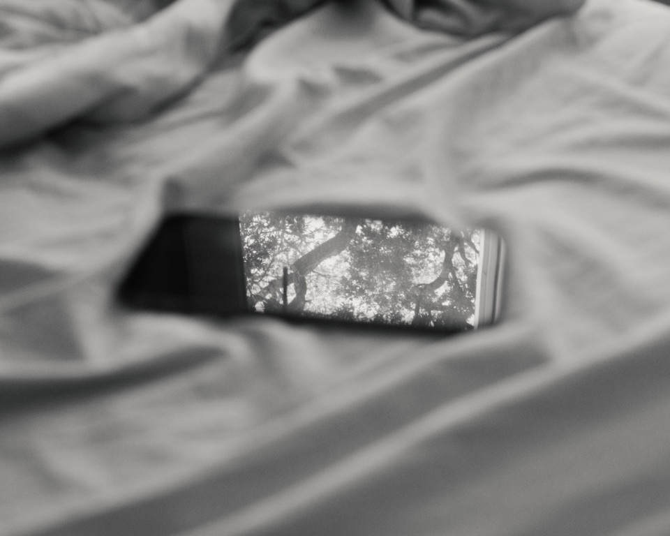 A phone on an unmade bed, its dark screen reflecting trees